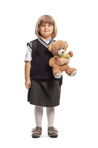 Full length profile shot of a little schoolgirl in a uniform holding a teddy bear isolated on white background