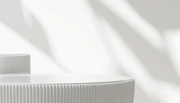 istock Two glossy white round cylinder ceramic podium  in corrugated side in sunlight and leaf shadow on white wall 1450229782