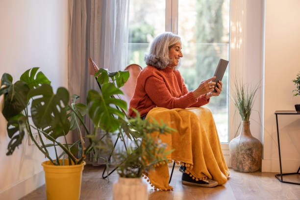 starsza kobieta z siwymi włosami, w salonie w domu siedząc w fotelu przy oknie, domu z wieloma roślinami i nowoczesną dekoracją, ma tablet do rozmowy wideo z rodziną - grandparent family reading inside of zdjęcia i obrazy z banku zdjęć