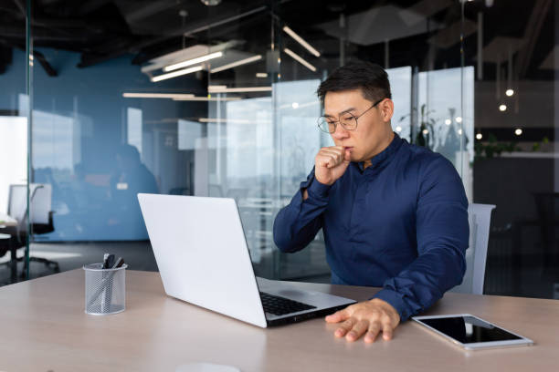 istock Asian man coughing at work in office, businessman working sick at workplace man using laptop, mature boss in glasses having strong cough 1450252600