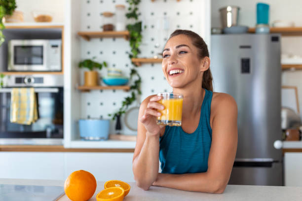 bella giovane donna che beve succo d'arancia fresco in cucina. dieta sana. giovane donna felice con un bicchiere di succo e arancia a tavola in cucina. - mature women foto e immagini stock