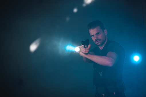 Young Caucasian policeman looking for a murderer in the darkness. He is wearing a bullet proof vest and holding a fire gun.