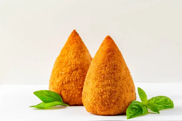 Italian rice balls that are stuffed, coated with breadcrumbs and deep fried.  Filled with ragù, mince meat, caciocavallo cheese and green peas. Conical-shaped arancini with basil on a white background. Sicilian food.
