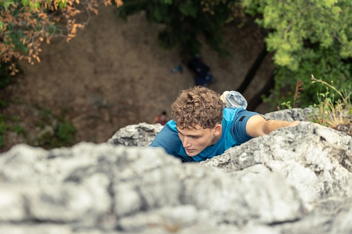 An overhead shot of a male with curly blond hair and in a blue T-shirt, climbing a cliff