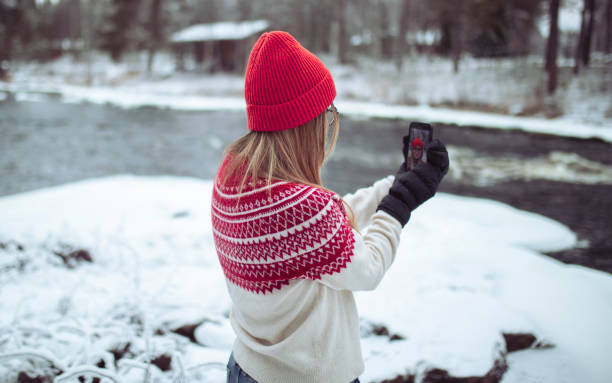 Back view of a woman in winter clothes stands on the coast and takes a selfie
