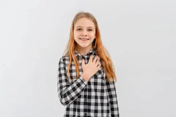 Smiling teen girl looks flattered, holds hand on chest, feels appreciation, stands in casual plaid shirt over white studio background