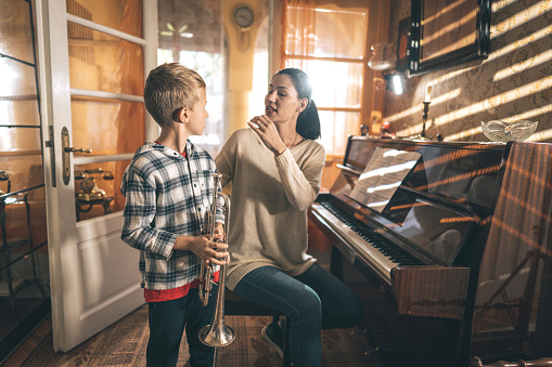 Young boy playing a trumpet, while his music teacher is showing him how to breathe while playing, they are together at home.