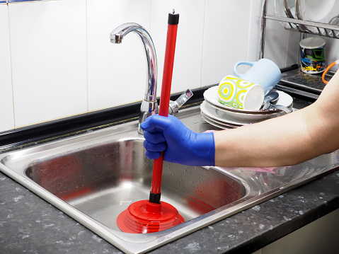 Removing blockage of the pipe, drain in the sink with a plunger. Stainless steel kitchen sink and red tools.