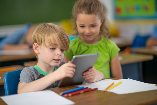 Two elementary school children work together on a tablet in class.  They are both dresed casually as they use the device to help facilitate their work.
