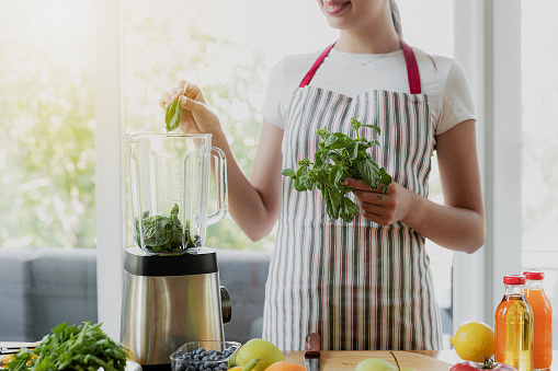 Young smiling woman cooking green spring smoothie at home kitchen using electronic blender device. Close up of beautiful happy girl hands put leaves into a gadget bowl for mixing. Ingredients on table