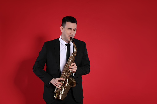 Young man in elegant suit playing saxophone on red background