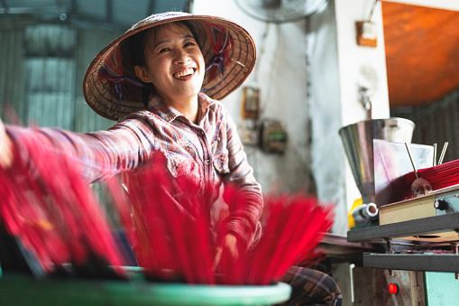 portrait of smiling vietnamese woman at work bench sorting red incense sticks in green bucket