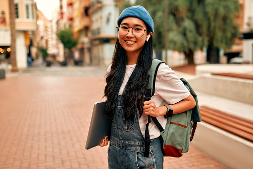 Portrait of asian hipster woman with backpack holding laptop, listening to music in wireless headphones, standing on city street. Asian student at the campus.