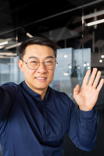 Vertical shot, Asian businessman talking on video call, man looking at smartphone camera and waving cheerfully, greeting gesture, programmer working inside office in glasses and shirt.