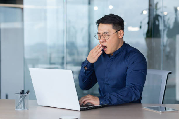 istock Tired worker at work yawning, asian man in shirt and glasses working inside office using laptop, man sleepless at work 1450339922