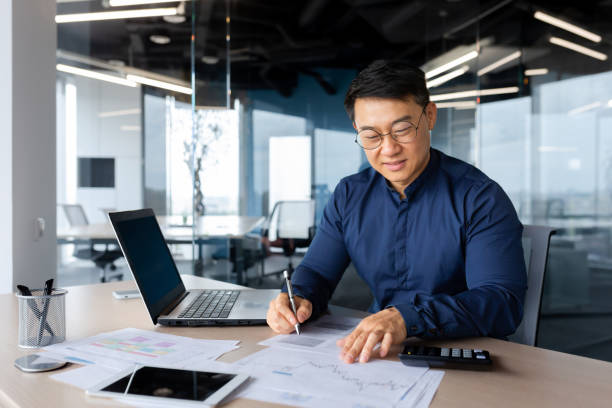 istock Successful and happy investor working with documents and charts inside office, Asian businessman behind paper work calculates bills with calculator, man at work with laptop happy with achievements 1450340089