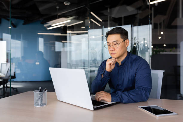 серьезно мыслящий азиатский бизнесмен, работающий в современном офисе, зрелый мужчина в рубашке и очках, использующий ноутбук на работе, ин - reflective workwear стоковые фото и изображения