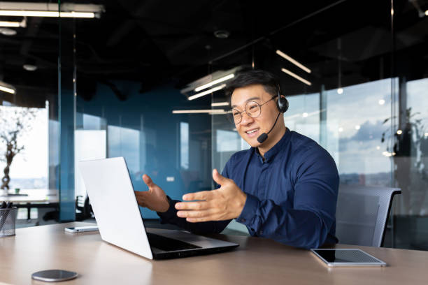 istock Asian with a headset is talking on video call, man inside office smiling and gesturing joyfully in an online meeting, a businessman is telling his colleagues, a customer service tech support worker 1450340247