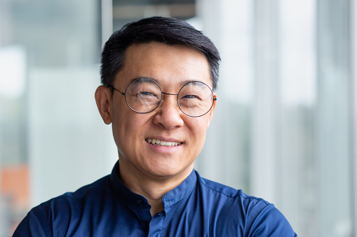 Close up photo portrait of successful and happy Asian businessman, mature boss with glasses working inside modern office building senior investor smiling and looking at camera