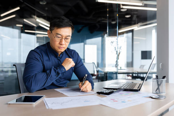 istock Serious thinking asian man working with documents inside office, concentrated businessman with contracts and bills reading and calculating reports, mature financier with documents 1450340319