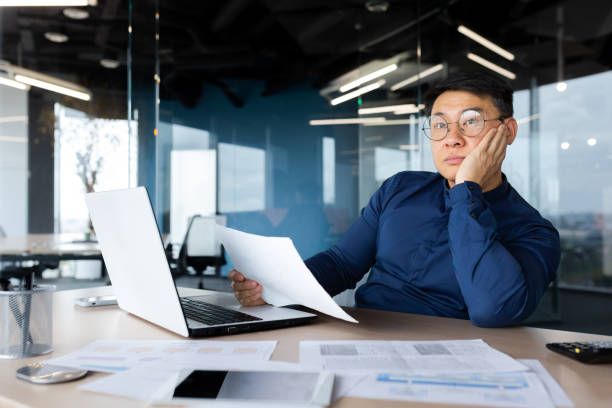 istock Serious thinking asian man working with documents inside office, concentrated businessman with contracts and bills reading and calculating reports, mature financier with documents 1450340322