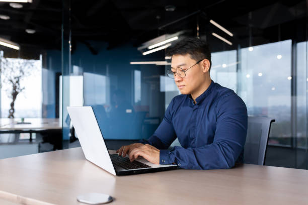 istock Serious and thoughtful businessman working inside office sitting at table using laptop at work, mature asian boss in shirt thinking and typing on keyboard 1450340449