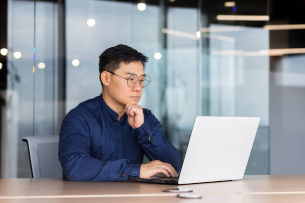 istock Serious and thoughtful businessman working inside office sitting at table using laptop at work, mature asian boss in shirt thinking and typing on keyboard 1450340463