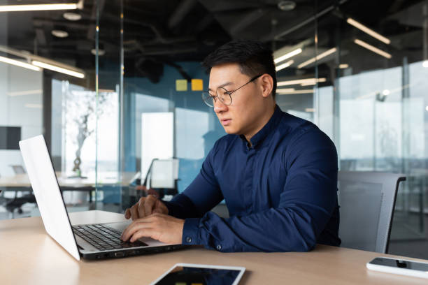 istock Serious and thoughtful businessman working inside office sitting at table using laptop at work, mature asian boss in shirt thinking and typing on keyboard 1450340465