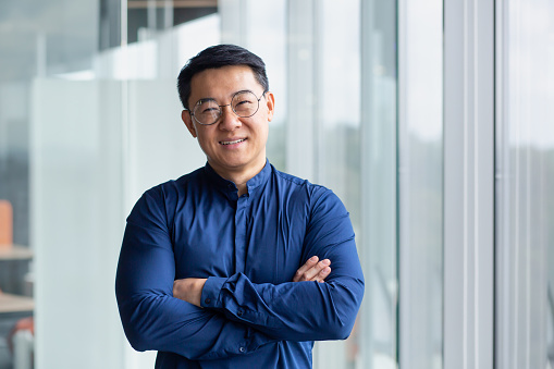 Portrait of successful mature boss, senior businessman in glasses Asian looking at camera and smiling, man with crossed arms working inside modern office building