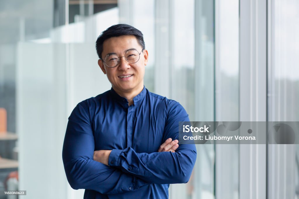 Portrait of successful mature boss, senior businessman in glasses Asian looking at camera and smiling, man with crossed arms working inside modern office building Portrait of successful mature boss, senior businessman in glasses Asian looking at camera and smiling, man with crossed arms working inside modern office building. Headshot Stock Photo