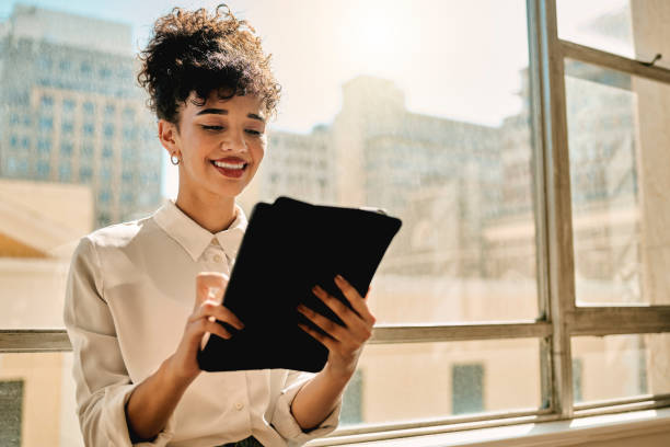 istock Business woman, digital tablet and relax against a window while planning, thinking and manage online task, happy and smile. Black woman, app and creative with internet search in office for ideas 1450341054