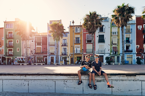 Two teenagers sightseeing beautiful town of Villajoyosa in Spain. 
They are sitting by the main promenade in front of the colorful houses.
Sunny summer day.
Canon R5