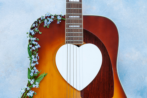 Acoustic guitar with beautiful blue spring forgot-me-not flowers and white heart shape silhouette on blue sky background.