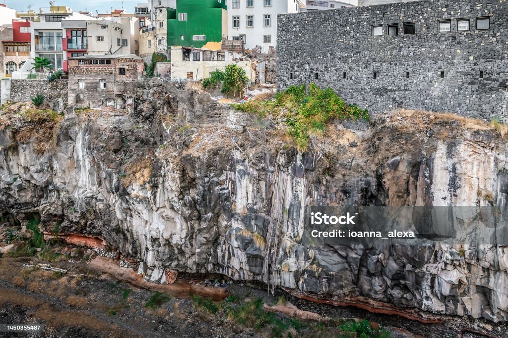 Barranco de Santos ravine with homeless dwellings at the bottom in Santa Cruz de Tenerife, Spain Barranco de Santos ravine with homeless dwellings at the bottom in Santa Cruz de Tenerife, Spain. City buildings on top of a cliff and tents made of blankets under a rock At The Bottom Of Stock Photo