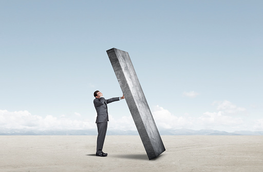 A businessman covers his eyes and extends his arm in a futile effort to prevent a heavy wall from falling on him.