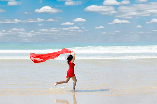 Young woman in red dress with red scarf running on beach in sunlight. Location: Arcachon, Atlantic Ocean, France