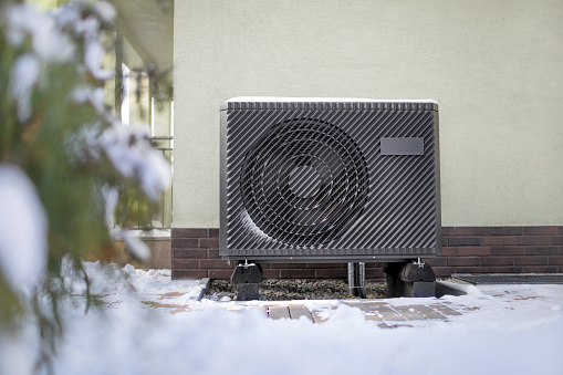 Photo of a modern air source heat pump unit standing against the residential building wall. Shot in winter, in a snowy setting.