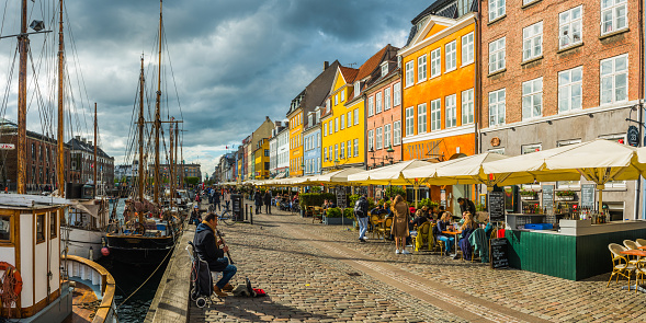 Crowds of people sitting, walking, chatting and relaxing on the picturesque waterfront of Nyhavn, its brightly coloured historic houses, cafes, bars, restaurants and nightclubs reflecting in the tranquil blue waters of the harbour canal leading to Kongens Nytor.