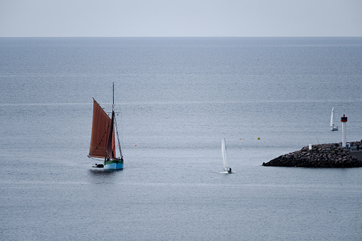 White sailboat floating on the sea surface by the rocky coastline with littoral vegetation at remote location, clear blue sky and island in the distance