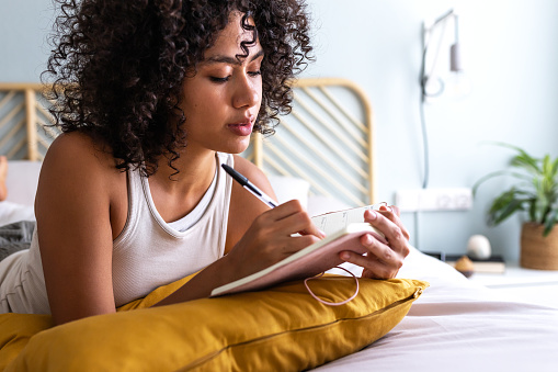 Pensive young multiracial latina woman lying down on bed with serious expression writing on journal in cozy bedroom. Lifestyle concept.