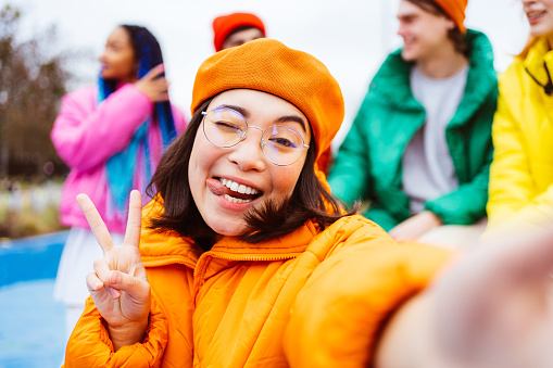 Multiracial group of young happy friends meeting outdoors in winter, wearing winter jackets and having fun, asian woman taking pov selfie - Multiethnic millennials bonding in a urban area, concepts about youth and social releationships