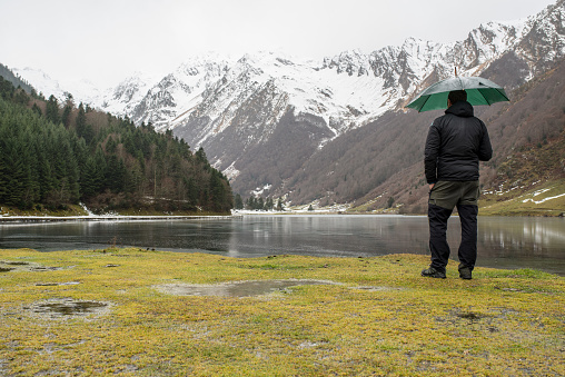 Rear view of a standing man holding an umbrella and looking at a snowy mountains at a lakeshore in French Pyrenees while is snowing.
