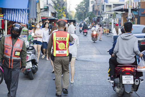 Thai traffic cop is using mobile phone and walking in street of residential district in Bangkok Chatuchak. In street are some  more people and a person is driving motorcycle