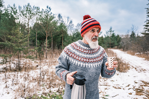 Christmas tree hunt in nature, Mature man with beard having a  warm beverage outdoors. He is wearing knit sweater and hat with jeans. Outdoor of the farm and wooded area in winter.
