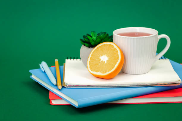 тетради, чашка чая и апельсин на зеленом фоне, пространство для копирования. - stationary pen orange work tool стоковые фото и изображения