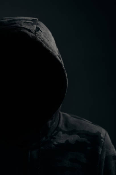 istock Man in dark camouflage hoodie with covered face dark background, hacker bandit and attacker. 1450398802
