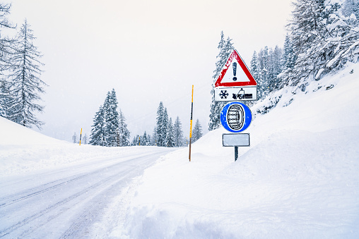 Warning and tyre chains signs along a snowy mountain road during a snowstorm in winter.