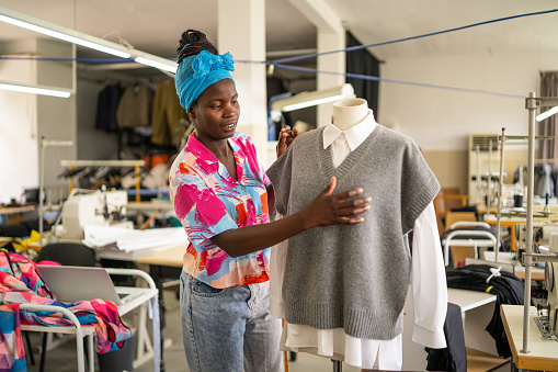Skilled experienced african-american fashion designer in a professional atelier standing in front of a mannequin with a tape measure on her neck, and making a new clothes