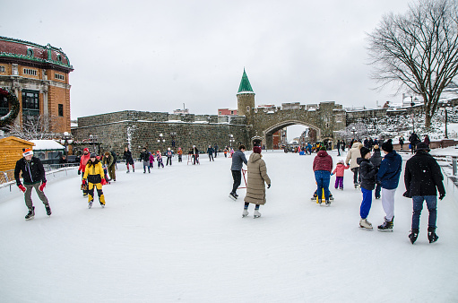 People and family ice skating on Place D'Youville municipal ice rink downtown Quebec city during winter day.\nThe St. John's gate is in background.