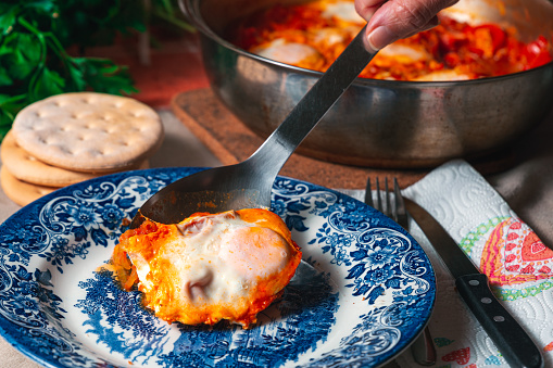 Close up view of a laddle pouring shakshuka on an empty plate, cooking pan, fresh parsley and gluten free bread